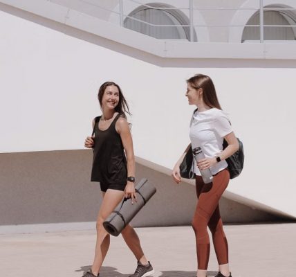Two women in active wear walking outdoors while smiling and carrying yoga mats.